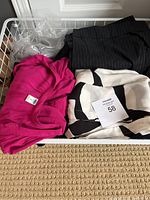 Wire basket filled with assorted women's clothing items including bright pink blouse, black and white patterned blouse, black pinstriped pants