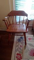 Photo showing one of the two wooden captain's chairs with curved backrest, vertical spindles, armrests, and turned legs, placed on a floral carpet near a window.
