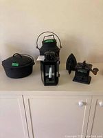 Full front view of all four items on white surface: cast iron pot with lid, iron jug, metal grinder, and metal lantern with candle