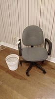 Grey upholstered office chair with black armrests and wheeled base next to white wicker waste paper basket on hard floor against striped wallpaper.
