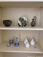Shelf with various decorative items: a brown vase with floral patterns, a decorative plate with green foliage on a stand, and an ancient Greek style brown vase.