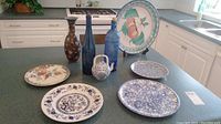 Overview of lot showing 5 plates, 3 tall bottles, cloisonné vase and small teapot on kitchen counter