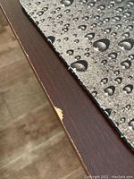Close-up photo of desk surface showing chipped veneer at the edge with a dark wood finish and textured desk mat on top with water droplet print.
