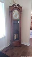Full front view of tall wooden grandfather clock with glass door and brass weights and pendulum visible.