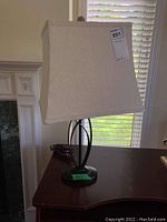 Table lamp showing full lamp with beige rectangular fabric shade and dark metal base on dark surface near window.