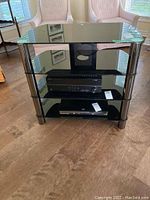 Front view of the media stand with three glass shelves and black metal cylindrical legs, set on wood flooring. Electronic devices shown are not included.
