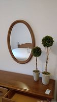 Oval wood framed mirror hanging on wall above wooden surface with two faux topiary plants in white pots beside it.