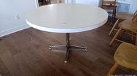 Full view of the round table with white laminate surface, showing some scratches and the metal base with four legs ending in white floor protectors.