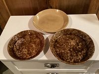 Three antique pottery pie plates grouped on white surface, two mottled brown and one beige yelloware plate