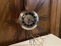 Front view of mid century modern sunburst electric wall clock with brass-tone and black metal rods and Roman numeral clock face