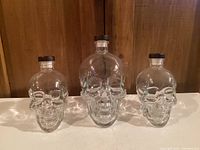 Front view showing three crystal skull-shaped vodka bottles of varying sizes on a white surface with wooden background.