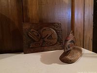 Overall view of the two wood carvings, one mounted relief and one 3D painted fish carving on base.