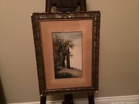 Full view of framed watercolour painting on easel, showing the artwork, mat, and ornate frame with some damage.