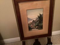 Full view of framed watercolour painting showing lakeside village with church and two figures.