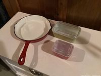 Enamel cast iron frying pan with two glass covered Pyrex dishes on white surface against wood background.