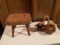 Milking stool and bird carving displayed side by side on a white surface with wood paneling background.