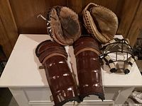 Photo showing two vintage leather baseball gloves, a pair of brown leather shin pads, and a catcher's mask arranged on a small white table against a wood-paneled wall.