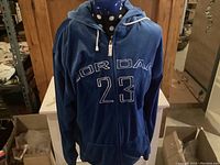 Front view of vintage Michael Jordan hoodie on a mannequin, displaying blue velour fabric with large 'JORDAN' and '23' embroidery across chest, zipper closure, and hood with white drawstrings.