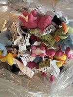Photo showing multiple small stuffed animal toys in assorted colors inside a transparent bag. Toys include dinosaur-like creatures with tags attached.
