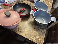 Four non-stick pots and pans in various colors and sizes placed on a table. Includes a frying pan with red handle, two blue handled pots, and one pink pot with glass lid.