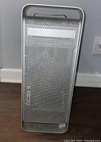 Front view of Apple desktop tower computer with perforated metal front panel including optical drive slot and multiple ports.