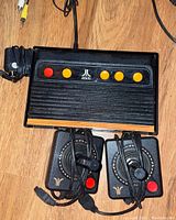 Atari console with wood grain front, four yellow buttons and two red buttons, two joystick controllers with red buttons and cables, power adapter on wooden surface.