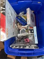 Overhead photo of blue plastic container filled with various boxed and wrapped new reseller items including HDX adjustable metal shelf, Duracell AA batteries, kitchen tools, and miscellaneous small household items.