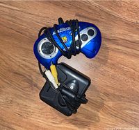 Blue Sega Genesis 16-bit controller with attached black cable alongside a black AC power adapter with its own cable, shown on a wood surface.