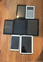 Photo showing six Android tablets arranged on wooden floor, including 2 Samsung, 2 Amazon, 1 Nook, and 1 Lenovo tablets. Some have cracked screens.