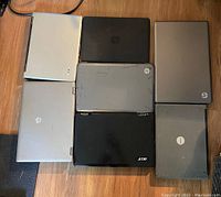 Top view showing all 7 laptops closed on a wooden floor. Includes 4 HP, 2 Acer, and 1 Dell laptop with visible wear and scratches.