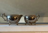 Mini silver tone creamer and sugar bowl set shown from the front on a mantel shelf.