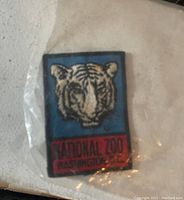 Photo of a rectangular embroidered cloth patch featuring a tiger face on blue background and red text banner below reading 'NATIONAL ZOO WASHINGTON D.C.'. Patch is sealed in clear plastic.