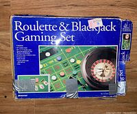 Outer blue box labeled "Roulette & Blackjack Gaming Set" with pictures of the roulette wheel and gaming board. Box shows condition wear and tape repairs.