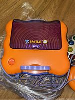Orange and purple VTech V Smile console top view showing V Smile logo and power button, with controller wires coiled.