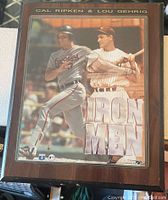 Wood plaque showing two baseball players Cal Ripken in Orioles uniform and Lou Gehrig in Yankees uniform framed with text 'Cal Ripken & Lou Gehrig' on top and 'IRON MEN' in large font below.