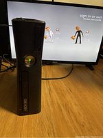 Photo of black Xbox 360 console standing vertically connected to a TV showing Xbox sign-in screen. The console's power button is illuminated. The console has no top cover and scratches.