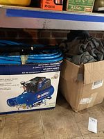Photo showing the box of Campbell Hausfeld 3 Gallon Air Compressor Kit with attached blue hose and a cardboard box with car covers.