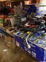 Photo showing various boxed automotive tools including Goodyear 6 Ton hydraulic jack, lug wrench, and jumper cables on a shelf.