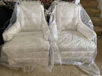 Two cream-colored jacquard upholstered armchairs covered in protective plastic, showing front view with floral pattern and button-tufted backs.