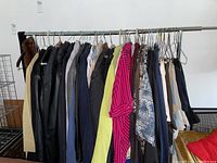 Full view of clothing rack with multiple women's garments including coats, shirts, and pants in various colors and fabrics, hung on metal hangers.