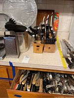 Photo showing two wooden knife blocks filled with assorted knives, metal cooking utensils, a box grater, and a masher on the kitchen counter with a partially open drawer of assorted cutlery below.