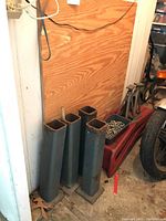 Four tall steel auto jack stands with square bases and square openings leaning against the wall with two metal auto ramps and one smaller jack stand visible nearby.