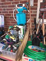 Wide view of assorted lawn and garden tools including sprayer, nozzles, hoses, and hand tools organized in containers and leaning against a brick wall.