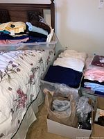 Photo of plastic containers filled with folded women's clothing including sweaters, T-shirts, and slacks beside bed and boxes with other items.