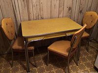 View of rectangular Arborite table in mustard yellow with three matching chairs exhibiting discoloration, set on linoleum patterned floor against wood paneled wall.