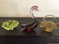 Three decorative glass items: lime green ashtray, purple-pink swan figurine, and amber basket arranged on a table.