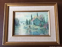 Framed art print of a cabin by a river with birds flying overhead, signed by Keirstead, in a gold and cream textured frame.