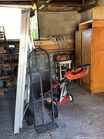 Photo of a folding aluminum ladder, black metal hand truck, and red Noma electric lawn mower with grass catcher in a garage.