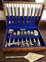 Overview of the complete flatware set in wooden storage box, showing knives, forks, spoons and serving utensils organized in blue velvet and cream satin lining.