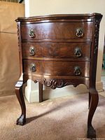 Front view of wood cabinet showing three drawers and decorative brass handles, curved legs with claw feet, and carved apron detail.